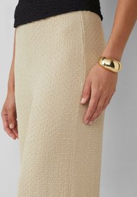 Beige textuurbroek met een geweven patroon, gecombineerd met een gladde, gepolijste gouden armband om een hand die op de dij rust.