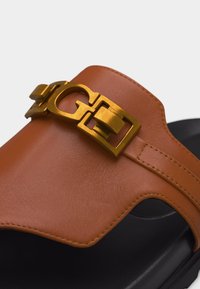 Καφέ δερμάτινο σανδάλι χωρίς λουράκια με μαύρη σόλα και χρυσή μεταλλική αγκράφα στη πάνω λωρίδα.