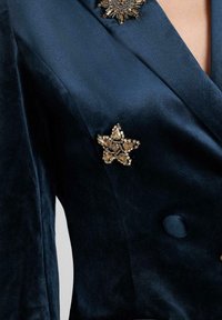 Marineblauwe satijnen blazer met een gouden juweelster-speld en een decoratieve bloemenpin. Soepele textuur met een gestructureerd ontwerp en knoopdetails.