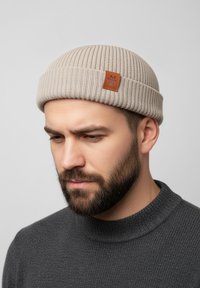 Beige ribbed knit beanie featuring a brown leather patch. Model is wearing a dark gray textured sweater, captured against a light background.