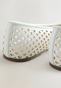Chaussures plates blanches avec un motif de découpes carrées et des semelles marron, montrant les détails latéraux et du talon sur un fond clair.
