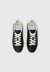 Baskets en cuir noir avec des lacets blancs et une semelle en caoutchouc crème. Présente un détail de logo doré sur le côté et un col rembourré.