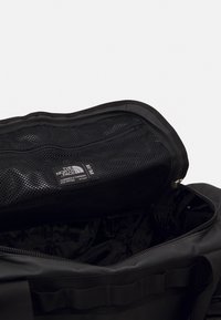 Sac de sport noir avec une poche en maille zippée, un compartiment intérieur à texture lisse, une étiquette imprimée et des détails de couture robustes.