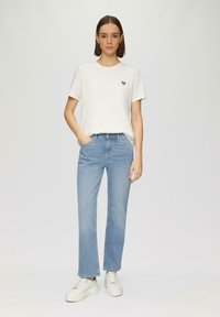 T-shirt en coton blanc avec un patch en forme de cœur bleu marine, associé à un pantalon droit bleu clair. La tenue est complétée par des chaussures blanches.