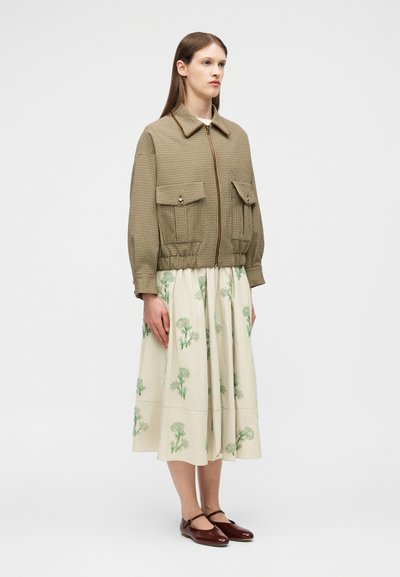 Femme portant une veste à fermeture éclair marron à carreaux, une jupe crème avec des imprimés floraux verts, et des chaussures Mary Jane marron, debout devant un fond uni.