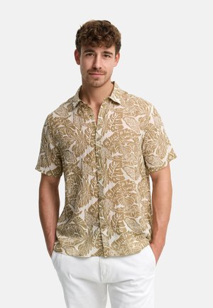 Ung mand med kort krøllet hår iført en beige og hvid kortærmet skjorte med tropisk print og hvide bukser, hænderne i lommerne.