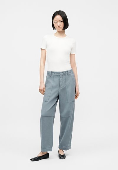 Γυναίκα με κοντά μαύρα μαλλιά που φοράει λευκό εφαρμοστό τοπ με κοντά μανίκια, άνετο μπλε παντελόνι με τσέπες-μπαλώματα και μαύρα επίπεδα παπούτσια, στέκεται μπροστά σε λευκό φόντο.