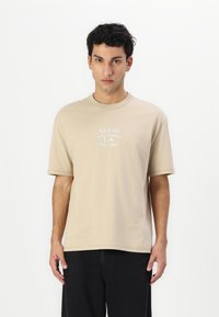 Beige bomull T-skjorte med korte ermer og rund hals. Har hvit trykt tekst: "GUESS CALIFORNIA L.A. Est. 1981."