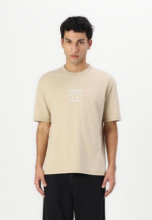 Beige cotton t-shirt with short sleeves and a crew neckline. Features white printed text: "GUESS CALIFORNIA L.A. Est. 1981."