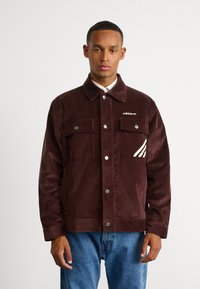 Burgundi värvi nõelkirjat jakk, millel on päästiknupud, kaks eespocketit ja valged Adidas triibud vasakul rinna taskul.