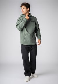 Grüner Fleece-Kapuzenpullover mit Kängurutasche und Kapuze mit Kordelzug, kombiniert mit schwarzer Jeans und weißen Sneakern, Modell mit Blick nach links.