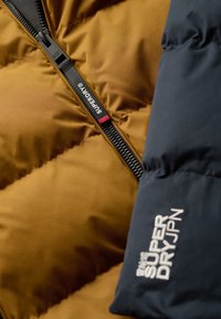 Brun pufferjakke med marineblå ærmer, der har en sort lynlås og et hvidt broderet logo "SUPERDRY" på ærmet. Glat tekstur, quiltet design.