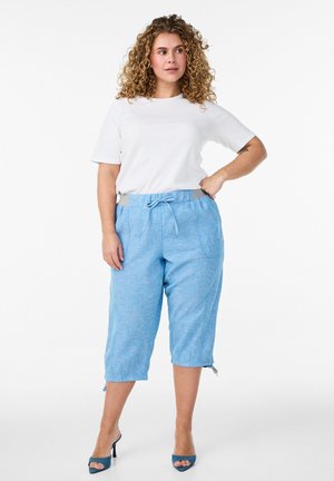 Kvinde med krøllet hår iført en hvid kortærmet skjorte, lyseblå korte bukser med snøre og blå sandaler med høje hæle, stående.