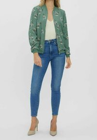 Zielona kurtka bomberka z kwiatowym nadrukiem, ściągacze na mankietach, zestawiona z jasnoniebieskimi obcisłymi dżinsami i beżowymi szpilkami. Tkanina o fakturze, prosty fason.