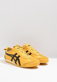 Paire de baskets basses jaunes et noires avec des lacets, présentée sur une surface blanche sur un fond gris clair uni.
