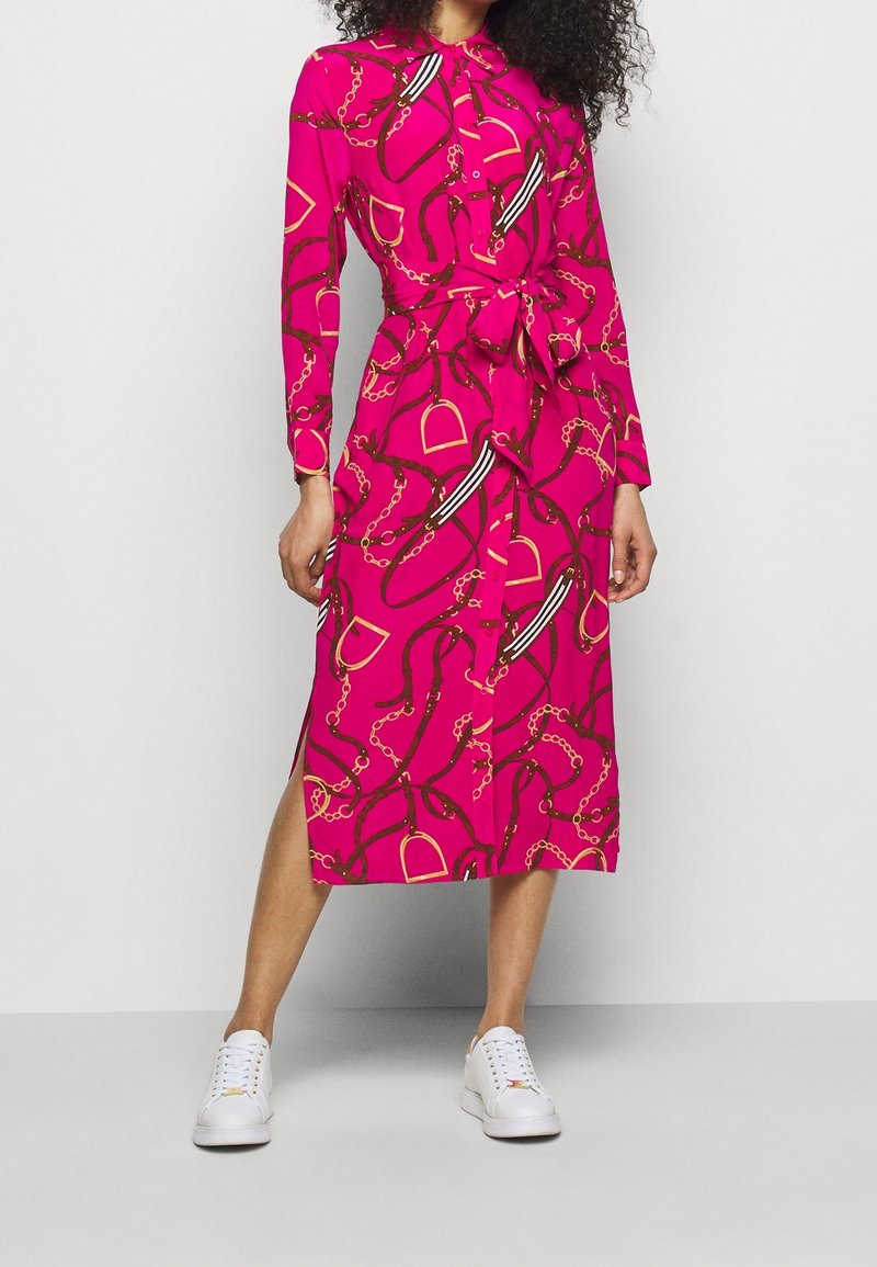 Kvinna klädd i en ljusrosa midiklänning med kedje- och bältesmönster, knuten vid midjan, matchad med vita sneakers mot en enfärgad bakgrund.