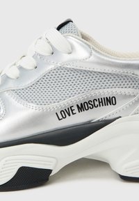 Srebrna tenisica s mrežastim panelima, bijelim vezicama, crnim naglascima i natpisom "LOVE MOSCHINO" sa strane. Teksturirana potplata za bolje prianjanje.