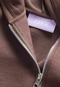 Felpa con zip marrone, caratterizzata da una zip argentata e un'etichetta del marchio lavanda con la scritta "JJXX". Tessuto morbido e testurizzato con una vista ravvicinata.