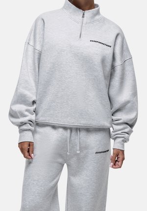 Osoba ubrana w jasnoszary dres z napisem „"Good For Nothing"” na piersi i udzie, stojąca na tle jednolitego, jasnego tła.
