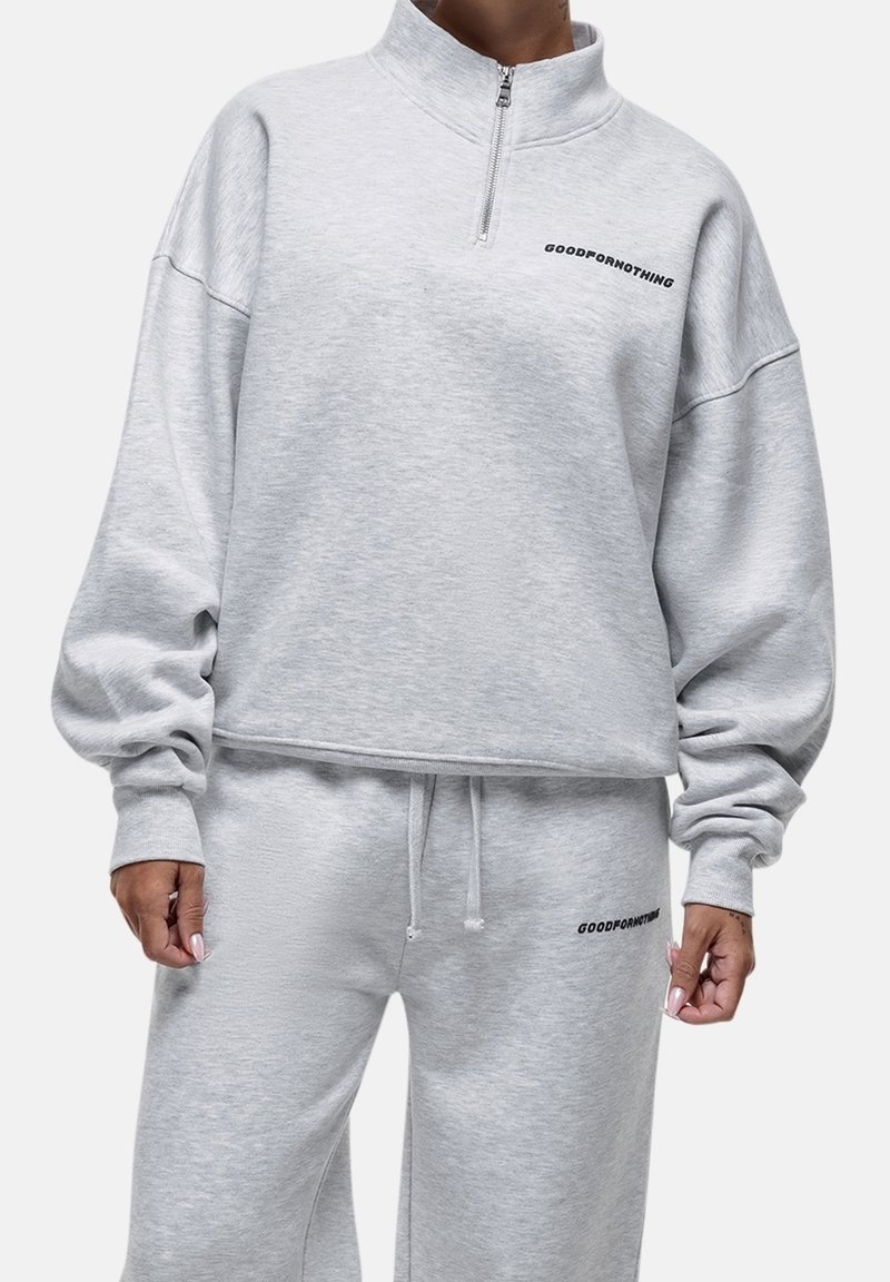 Personne portant un survêtement gris clair avec le texte « "Good For Nothing" » sur la poitrine et la cuisse, debout devant un fond uni clair.
