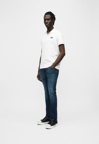 Άσπρο πόλο μπλουζάκι με κοντά μανίκια, σκούρο μπλε τζιν και μαύρα αθλητικά παπούτσια με λευκές σόλες. Το μοντέλο στέκεται σε ουδέτερο φόντο.