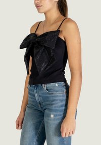 Zwart mouwloos topje met een grote, gestructureerde strik aan de voorkant. Gecombineerd met lichtblauwe, versleten jeans. Eenvoudig bandjesontwerp.