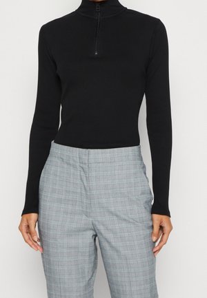 Haut noir à manches longues et ajusté avec un col zippé à l'avant, associé à un pantalon taille haute gris à carreaux porté par une personne debout.