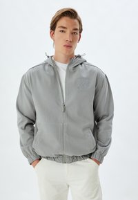 Chaqueta de color gris claro con cierre tipo cremallera, capucha, puños y dobladillo elásticos. Incluye dos bolsillos frontales y un sutil detalle de logo bordado.