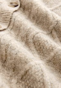 Beżowy sweter dziany o miękkiej fakturze, z prążkowanymi detalami i dwoma dużymi guzikami. Zbliżenie pokazujące głębię materiału i wzór.