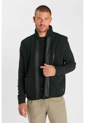 Uomo con capelli corti che indossa una giacca in pile nera con cerniera e pantaloni beige, tenendo con una mano la zona della cerniera della giacca.