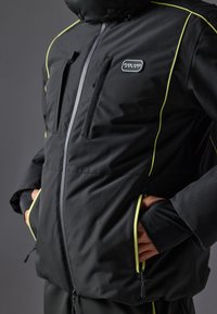 Chaqueta aislante negra con acentos amarillos, cremallera frontal, dos bolsillos con cremallera y parche de logo en el pecho. Cuenta con un alto cuello y puños ajustables.