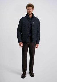 Marineblauwe gewatteerde jas met hoge kraag, ritszakken en een chevron-stiksels patroon, gecombineerd met donkere broeken en gepolijste schoenen.