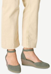 Zapatos de cuña verdes de lona con una suela intermedia trenzada, diseño de talón abierto y tira en el tobillo, combinados con pantalones tapering de color claro.