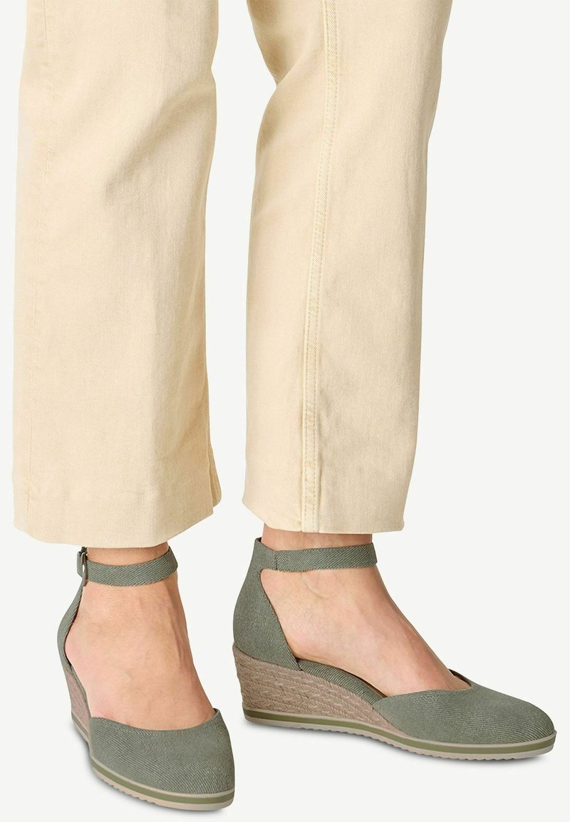 Zapatos de cuña verdes de lona con una suela intermedia trenzada, diseño de talón abierto y tira en el tobillo, combinados con pantalones tapering de color claro.