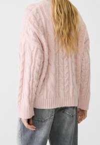 Sweter warkoczowy w jasnoróżowym kolorze z prążkowanymi mankietami i dołem. Wykonany z miękkiej przędzy, ma teksturowane wzory i luźny krój. Noszony z szarymi dżinsami.