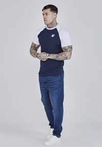 Námornícky modré tričko s bielymi raglánovými rukávmi, na hrudi so malým logom, v kombinácii s tmavomodrými džínsami a bielymi teniskami.