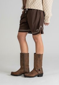 Shorts atletici din plasă maro cu accente albe și fante laterale, asortați cu cizmulițe din piele maro, până la genunchi, care au o fermoar lateral și detalii cu cataramă.