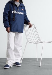 Granatowa kurtka przeciwwiatrowa z białym logo "ellesse" i regulowanym kapturem. W zestawie z białymi spodniami o szerokiej nogawce i granatowymi trampkami. W pobliżu minimalistyczne krzesło na metalowej podstawie.