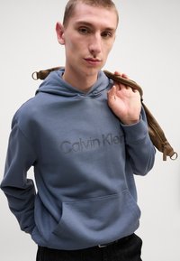 Zila kokvilnas kapuce ar kangaroo kabatu un izšūtu "Calvin Klein" logo. Modelis tur brūnu siksnu pār vienu plecu.