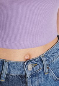 Lila ribbad crop top i kombination med slitna blå jeans. Toppen har en rak kant, medan jeansen har en metallknappstängning och gula sömmar.