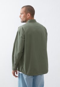 Olive green lightweight jacket with a relaxed fit, featuring a collared neck, long sleeves, and snap-button cuffs. Worn over a white shirt.