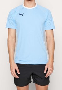 Camiseta atlética de manga corta color azul claro con detalle de cuello blanco y logotipo oscuro. Usada con pantalones cortos negros. Tela suave y ligera.