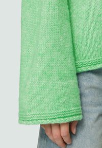 Grüner gestrickter Pullover mit weiten Ärmeln, der ein strukturiertes Muster und gerippte Bündchen aufweist. Über leichtem Denim-Stoff getragen.