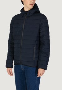Giacca puffer navy con cappuccio, caratterizzata da una texture trapuntata, chiusura frontale con zip e due tasche laterali. Design leggero e isolato.