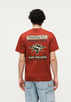 Mand med krøllet hår har en rød t-shirt på med teksten "Replay Inc Engineered Works" og grafiske pile, der omgiver en vinylplade på bagsiden.