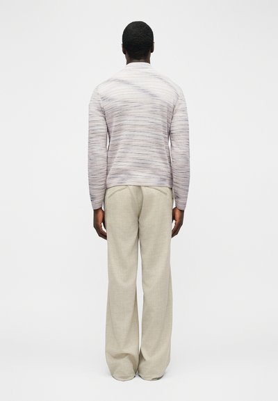 Homme debout, tourné de dos, portant un haut à manches longues gris clair texturé et un pantalon ample beige à jambes larges, sur un fond uni.