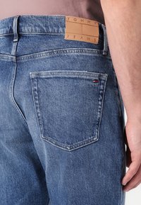 Jeans de mezclilla en azul claro con un acabado texturizado, que presentan una etiqueta de cuero marrón claro etiquetada "TOMMY JEANS" en la cintura trasera y costuras en contraste.