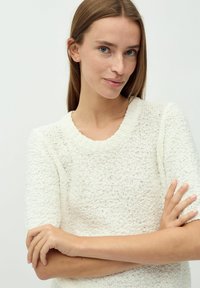 Sweter z dzianiny w kremowym kolorze, o okrągłym dekolcie i krótkich rękawach. Model ma ręce skrzyżowane, eksponując szczegóły odzieży.