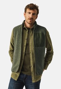 Green fleece vest with a zip closure, textured front, and a solid green pocket. Worn over a green button-up shirt, paired with dark jeans.