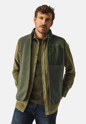 Green fleece vest with a zip closure, textured front, and a solid green pocket. Worn over a green button-up shirt, paired with dark jeans.